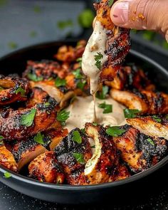 a person dipping sauce on top of chicken wings in a black bowl with green garnish