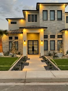 the front entrance to a modern home with lights on it's windows and landscaping