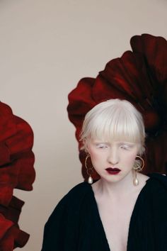 a woman with white hair and black dress standing in front of large red flower petals