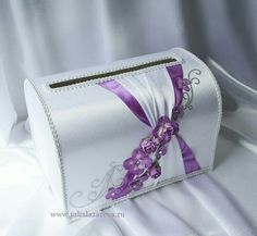 a white purse with purple ribbon and flowers on the front is sitting on a bed