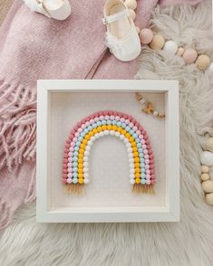 a white frame with a rainbow beaded decoration in it and some baby shoes on the floor