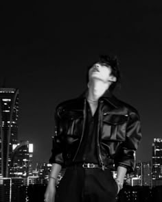 a man standing in front of a city skyline at night with his eyes closed, wearing a leather jacket