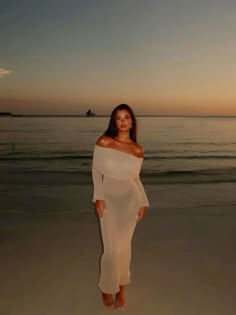 a woman standing on top of a sandy beach next to the ocean at sunset with her hands in her pockets