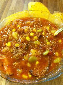 a glass bowl filled with chili and corn on top of a wooden table next to a piece of bread