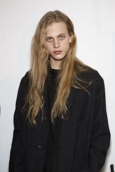 a woman with long blonde hair standing in front of a white wall wearing a black coat