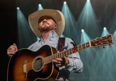 a man wearing a cowboy hat and holding an acoustic guitar in front of stage lights