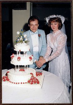 a man and woman standing next to each other in front of a white wedding cake