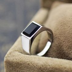 a silver ring with a square black stone on the center sits on a tan cushion