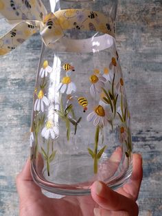 a hand holding a glass vase with daisies and bees painted on the side,