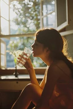 a woman sitting in front of a window drinking from a glass with the sun shining through it