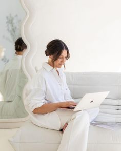 a woman sitting on a white couch using a laptop computer while looking at her reflection in the mirror