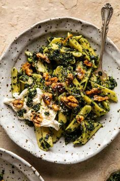 a white bowl filled with pasta covered in pesto and cheese next to two silver spoons