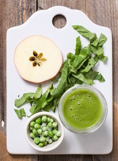 green smoothie ingredients displayed on white surface, including apple and leafy greens in small bowls