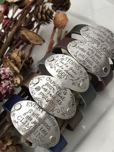 four metal bracelets with writing on them sitting next to dried flowers and pine cones