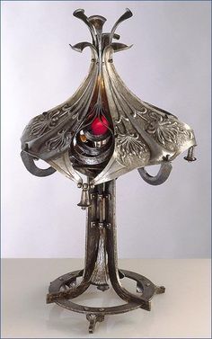 an ornate silver vase with red glass in the center on a metal stand against a white background