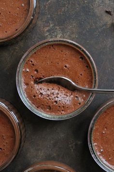 four jars filled with chocolate pudding on top of a metal counter topped with a spoon