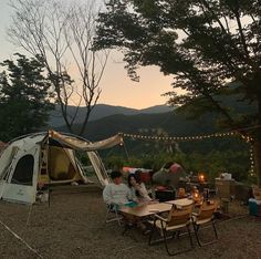 people sitting around a table in front of a tent with lights strung from the trees