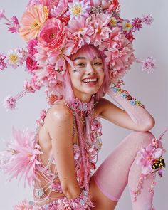 a woman with pink hair and flowers on her head, sitting in front of a white background