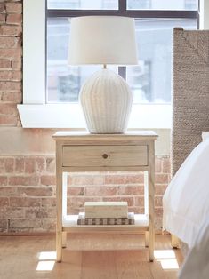 a white lamp sitting on top of a wooden table next to a bed in front of a window