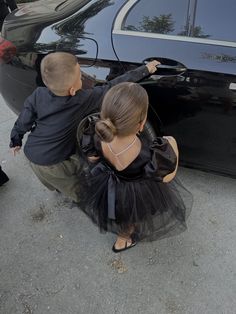 two children are standing next to a black car and looking at something on the ground