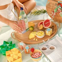 a woman sitting on a bed with fruit and drinks in front of her, next to an open book