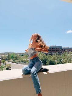 a woman sitting on top of a wall next to a tree filled sky scrapes her hair in the wind