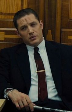 a man wearing a suit and tie sitting in a chair with his hands on his hips