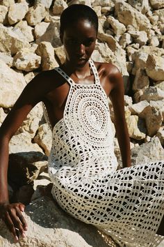 a woman sitting on top of a pile of rocks wearing a white crocheted dress