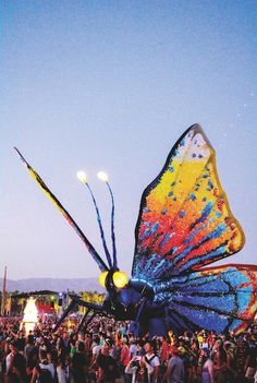 a large butterfly kite flying over a crowd of people at an outdoor event in the sun