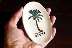 a hand holding an oval dish with a palm tree on it and the word aloha painted on it