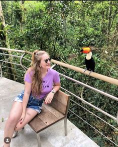 a woman is sitting on a bench looking at a toucan perched on the railing