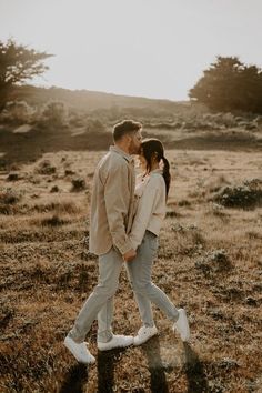 a man and woman are walking through the grass together in an open field at sunset