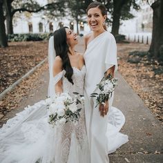 two brides standing together in front of some trees and leaves on the ground with one holding her bouquet