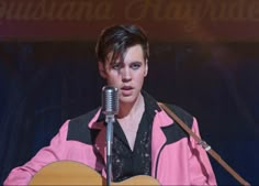 a young man holding a guitar while standing in front of a microphone and wearing a pink jacket