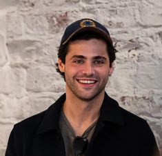 a smiling man wearing a hat and jacket in front of a white brick wall,