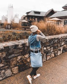 a woman wearing a denim vest and hat standing next to a stone wall holding a blue purse