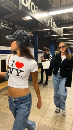 two women in jeans and baseball caps are walking through the subway station, one is wearing a t - shirt that says i love ny