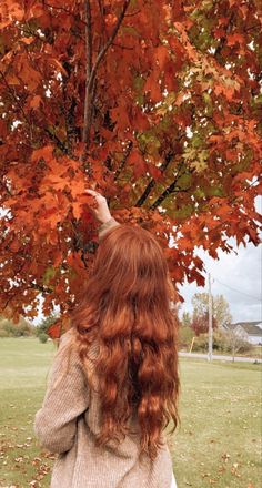 a woman with long red hair standing under a tree in the fall, reaching up for leaves