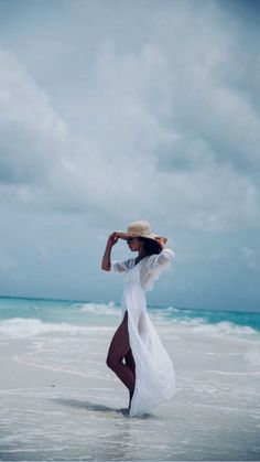 a woman in a white dress and hat walking on the beach with her arms behind her head