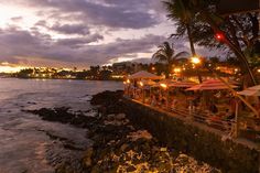 people are sitting at tables on the edge of the water as the sun goes down