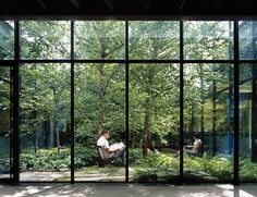 a man sitting in a chair looking out the window at some trees and people outside