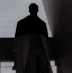 a man standing in front of stairs with his back turned to the camera, wearing a long coat