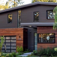 a modern house with wood siding and black garage doors on the front, surrounded by greenery