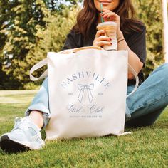 a woman sitting on the grass holding a tote bag with a drink in her hand