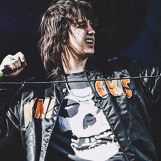 a man with long hair wearing a leather jacket and holding a microphone in his right hand