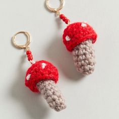 two crocheted mushrooms hanging from hooks on a white surface, one is red and the other is gray