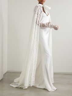 a woman in a white wedding gown and long cape with lace on the sleeves is standing against a wall