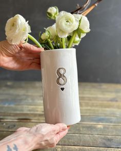 a person holding a white vase with flowers in it on a wooden table next to a wall