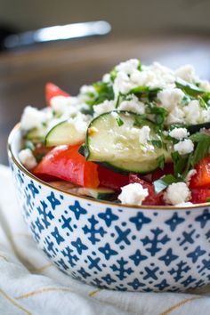 a blue and white bowl filled with cucumbers, tomatoes, feta cheese and herbs