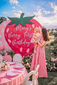 a woman holding a baby in front of a strawberry shaped sign that says merry first birthday